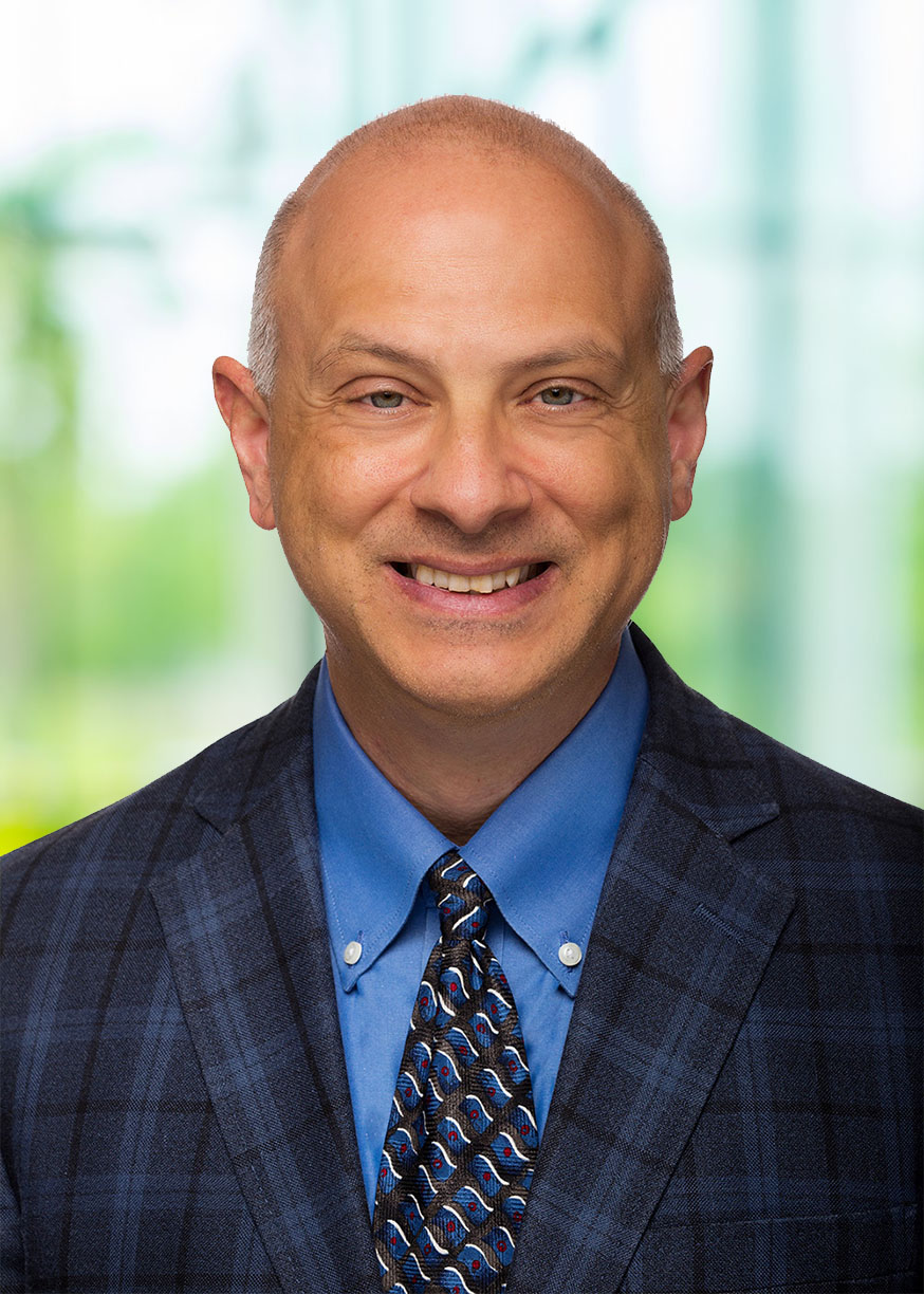 Smiling man wearing a blue dress shirt, patterned tie, and dark checkered blazer in front of a bright blurred background.